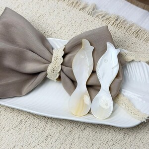 May include: Two mother-of-pearl spoons shaped like fish rest on a white, oval-shaped plate. A brown napkin is held by a lace napkin ring. The background includes a beige woven placemat and a decorative element.