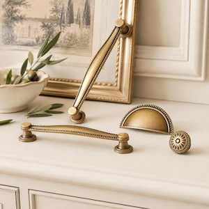 May include: A collection of antique brass hardware including two long arched drawer pulls with beaded edges, one semi-circular cup pull with a beaded border, and one round knob featuring a central floral petal design with a detailed beaded rim.
