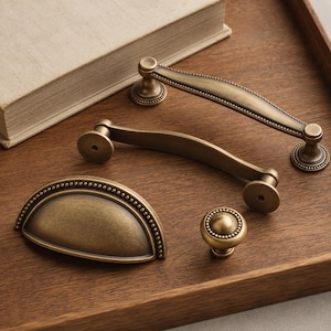 May include: A collection of four antique brass-toned metal cabinet hardware pieces, including two arched drawer pulls with beaded detailing, one semi-circular cup pull with a beaded edge, and one small round knob with a beaded border.