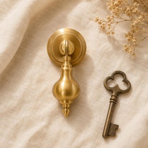 May include: A brushed brass drop pull handle with a circular backplate and a teardrop-shaped pendant, accompanied by a separate antique-style metal skeleton key with a clover-shaped bow and a rectangular bit at the end of the shaft.