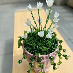 Puede incluir: Un arreglo floral artesanal con flores blancas en forma de tulipán, tallos y hojas verdes, en una maceta rosa. El arreglo incluye enredaderas de cuentas verdes y se encuentra sobre una superficie de madera marrón claro. Las flores son de un material translúcido.