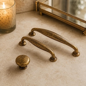 May include: Two arched cabinet pulls and one round cabinet knob, all featuring a textured, antique brass finish. The hardware pieces have a weathered, metallic appearance with dark brown and gold tones, showcasing a curved design for furniture.