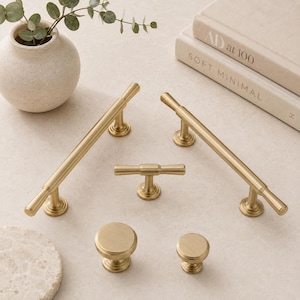 May include: A collection of five brushed brass cabinet hardware pieces including two long bar pulls, one T-shaped pull, and two round knobs. The hardware features a smooth, metallic finish with circular base plates and cylindrical handles.