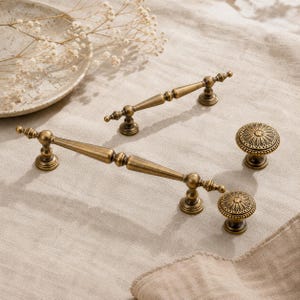 May include: Two antique brass cabinet pulls with a tapered, spindle-like design and two matching round cabinet knobs featuring a detailed, embossed floral pattern on the top surface. The hardware has a smooth, metallic finish with dark accents.