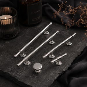 May include: A collection of polished silver-toned metal cabinet hardware, including three long bar pulls of varying lengths, one smaller T-bar pull, and one round knob. Each piece features a smooth, reflective finish and cylindrical design.