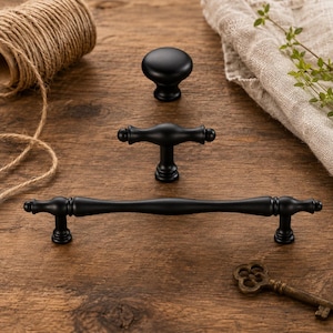 May include: Three matte black cabinet hardware pieces including one round knob, one small T-shaped pull, and one long horizontal handle. Each piece features a smooth, solid finish with rounded ends and a consistent dark, non-reflective surface.