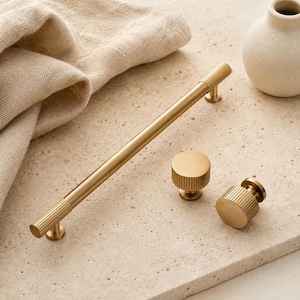 May include: A long, cylindrical gold-toned metal cabinet pull handle with ribbed detailing at both ends, accompanied by two matching circular cabinet knobs featuring a similar ribbed texture around their edges, all finished in a brushed metallic gold.