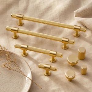 May include: A collection of gold-toned cabinet hardware including three long bar pulls with knurled textures, one T-bar knob, two round knobs, and one small cylindrical knob, all featuring a consistent brushed metallic finish and industrial design.