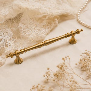 Peut inclure: Une poignée de meuble en métal doré brossé dotée d'une barre centrale effilée et fine avec des accents arrondis décoratifs et des bases de montage évasées à chaque extrémité, présentant une finition mate lisse et un design symétrique.