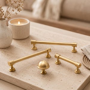 Può includere: Un set di quattro componenti per mobili in oro spazzolato, composto da tre maniglie cilindriche di diverse lunghezze e un pomello rotondo a forma di fungo, tutti caratterizzati da una finitura metallica liscia e linee pulite.