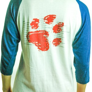 May include: A white and blue baseball style t-shirt with a red splatter design on the back. The shirt has a list of cities and dates printed on the back.
