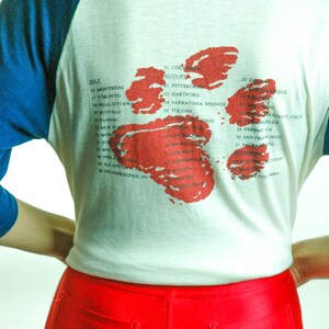 May include: A white t-shirt with a blue baseball sleeve design. The back of the shirt features a red and white map of the United States with a list of cities and dates. The text on the shirt reads: "July 16 Montreal, 17 Toronto, 18 Hull/Ottawa, 19 Buffalo, 21 Passaic, 22 New York, 23 Worcester, 24 Poughkeepsie, NY, 25 Boston, 26 Philadelphia, 27 Rochester, 28 Allentown, PA, 29 Poughkeepsie, NY, 31 Columbia, August 01 Pittsburgh, 03 Hartford, 04 Saratoga Springs, 05 Toledo, 20 Dallas, 21 Albuquerque, 22 San Diego, 23 Hollywood/Calif, 24 Fresno, CA, 25 Los Angeles, 26 San Francisco, 27 Sacramento, 28 Eugene, Oregon, 29 Seattle, 30 San Diego, 31  Washington, DC, 01  British Columbia"