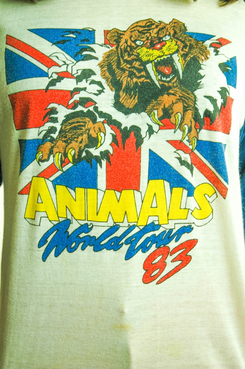 May include: A white t-shirt with a blue, red, and white Union Jack flag design. A brown and yellow tiger with a large fang is ripping through the flag. The text "ANIMALS World Tour 83" is printed below the tiger.