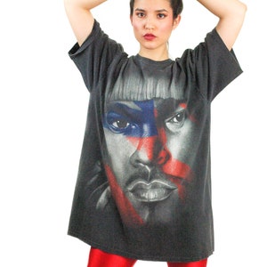 May include: A black t-shirt with a graphic print of a person's face. The face is partially obscured by red, white, and blue stripes. The person is wearing a black shirt and red pants.