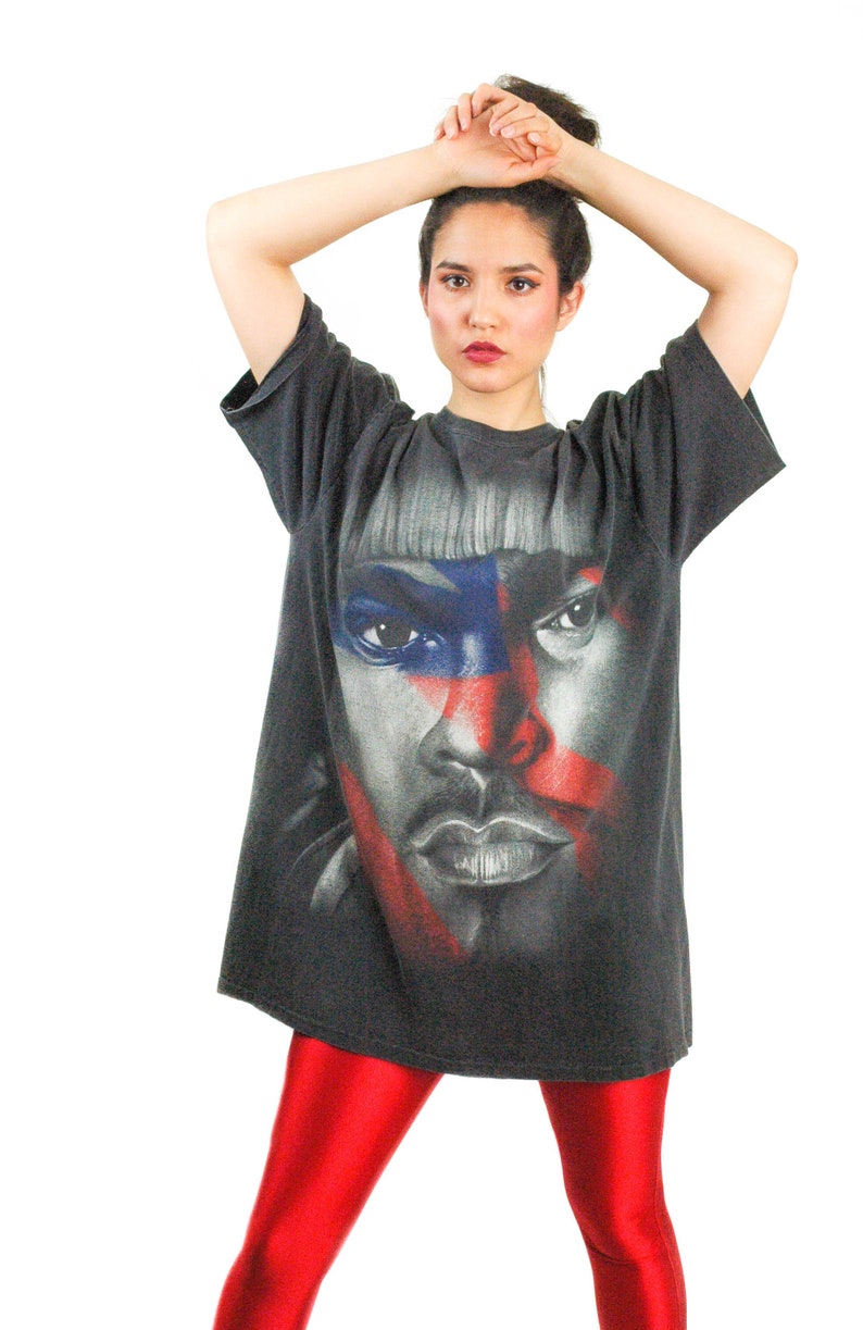 May include: A black t-shirt with a graphic print of a person's face. The face is partially obscured by red, white, and blue stripes. The person is wearing a black shirt and red pants.