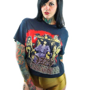 May include: A woman wearing a black t-shirt with a red, white, and black graphic of Ozzy Osbourne. The shirt has the text "Ozzy Osbourne" printed on the front. The woman has black hair and is wearing gold shorts.