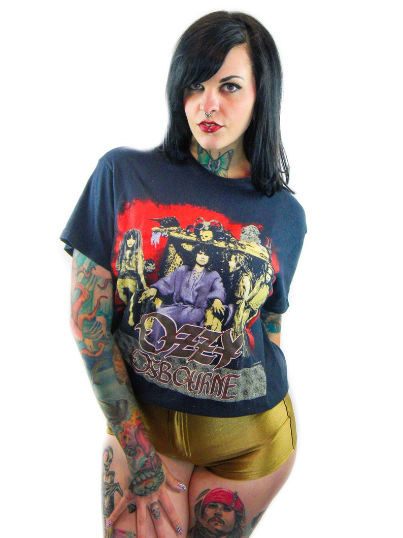 May include: A woman wearing a black t-shirt with a red, white, and black graphic of Ozzy Osbourne. The shirt has the text "Ozzy Osbourne" printed on the front. The woman has black hair and is wearing gold shorts.
