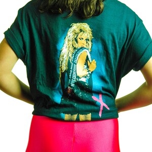 Puede incluir: Una camiseta verde oscuro con un estampado gr&aacute;fico de una mujer con el pelo largo y rubio que lleva un atuendo negro y un cintur&oacute;n plateado. La camiseta tiene una X rosa en la esquina inferior derecha.