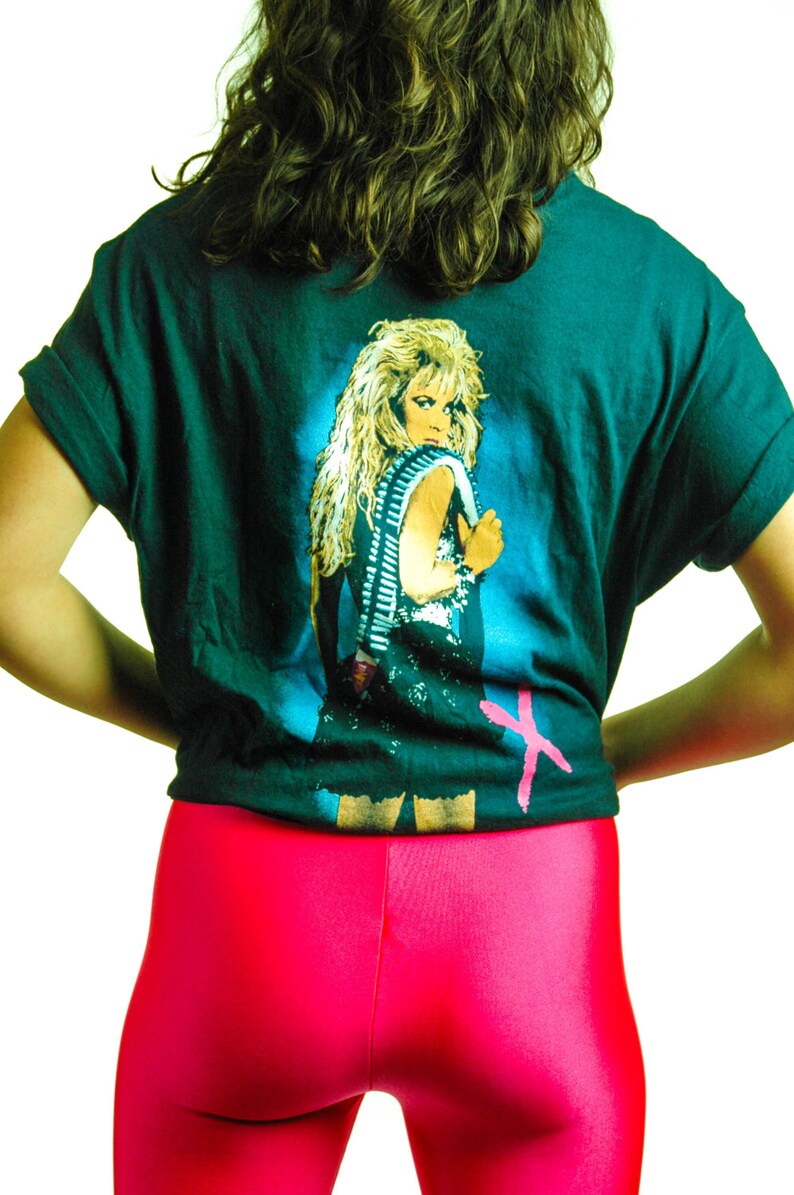 Puede incluir: Una camiseta verde oscuro con un estampado gr&aacute;fico de una mujer con el pelo largo y rubio que lleva un atuendo negro y un cintur&oacute;n plateado. La camiseta tiene una X rosa en la esquina inferior derecha.