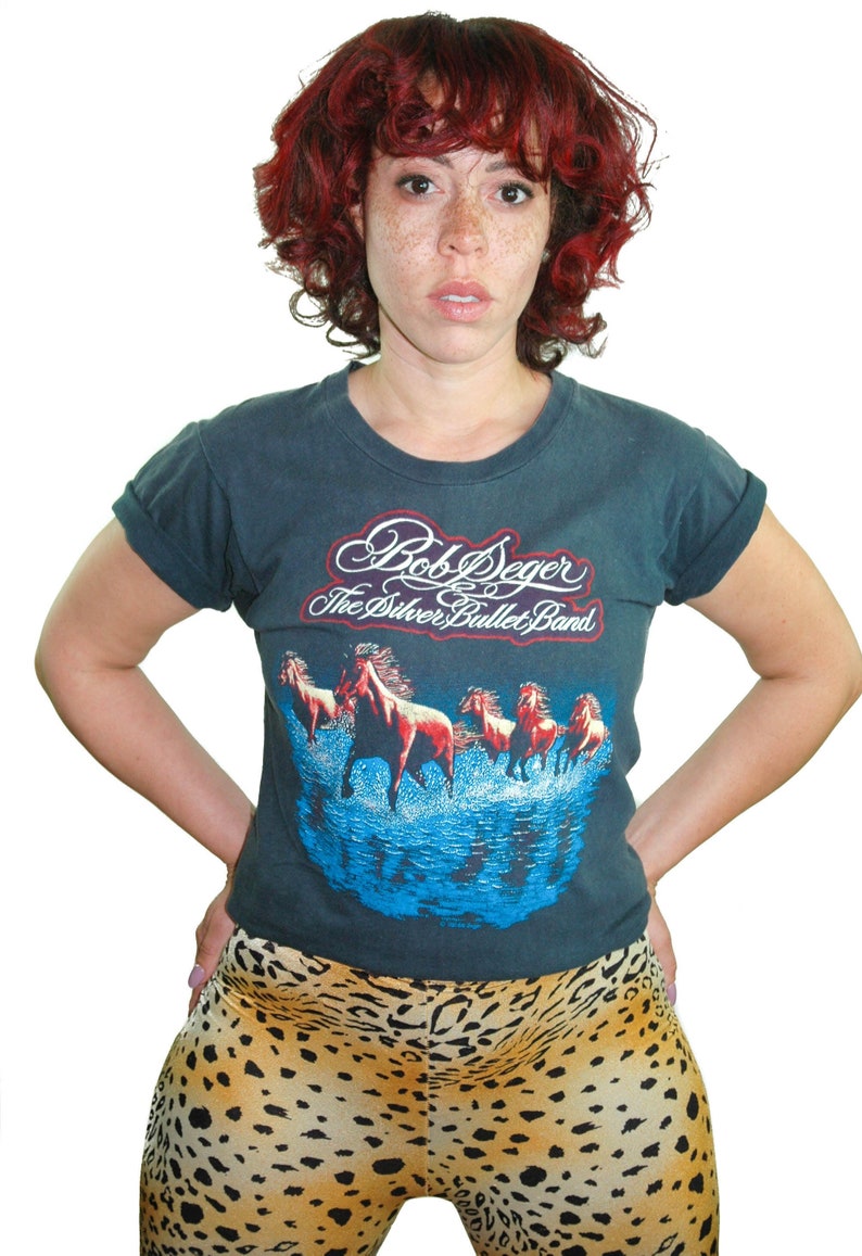 May include: A woman with red curly hair is wearing a dark blue t-shirt with a vintage Bob Seger and the Silver Bullet Band graphic. She is also wearing leopard print pants.