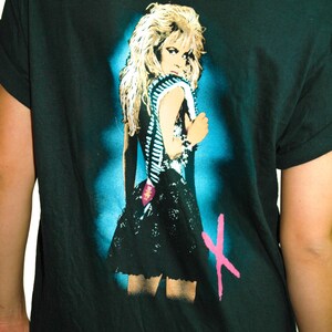 Puede incluir: Camiseta negra con un estampado gr&aacute;fico de una mujer con cabello rubio que lleva un vestido negro y un cintur&oacute;n tachonado. El gr&aacute;fico est&aacute; en un esquema de color azul y verde con una "X" rosa en el fondo.
