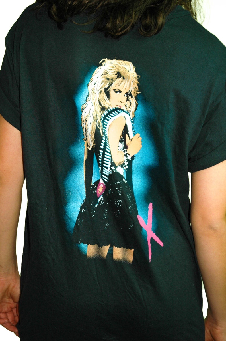 Puede incluir: Camiseta negra con un estampado gr&aacute;fico de una mujer con cabello rubio que lleva un vestido negro y un cintur&oacute;n tachonado. El gr&aacute;fico est&aacute; en un esquema de color azul y verde con una "X" rosa en el fondo.
