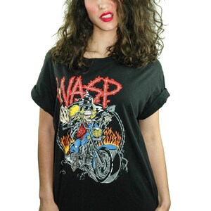 May include: Black t-shirt featuring a graphic of a cartoon character riding a motorbike with flames. The text "WASP" is printed in red on the shirt.