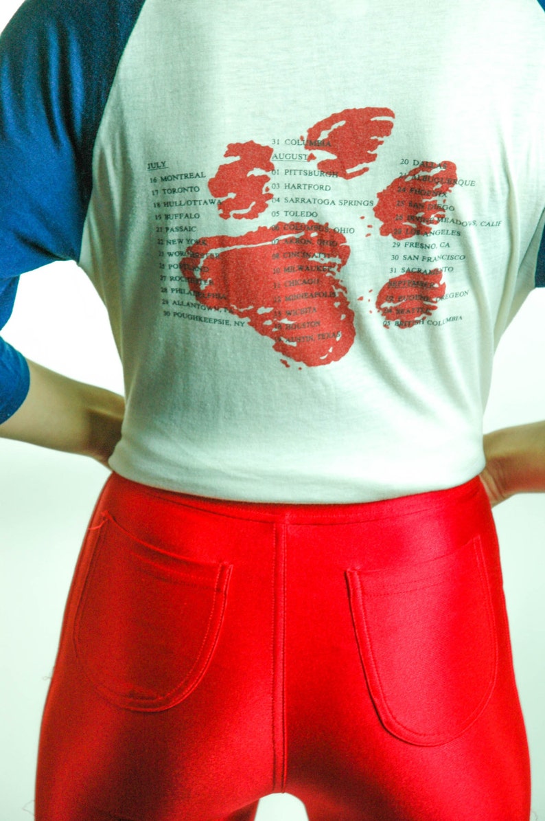 May include: A white t-shirt with a blue baseball sleeve design. The back of the shirt features a red and white map of the United States with a list of cities and dates. The text on the shirt reads: "July 16 Montreal, 17 Toronto, 18 Hull/Ottawa, 19 Buffalo, 21 Passaic, 22 New York, 23 Worcester, 24 Poughkeepsie, NY, 25 Boston, 26 Philadelphia, 27 Rochester, 28 Allentown, PA, 29 Poughkeepsie, NY, 31 Columbia, August 01 Pittsburgh, 03 Hartford, 04 Saratoga Springs, 05 Toledo, 20 Dallas, 21 Albuquerque, 22 San Diego, 23 Hollywood/Calif, 24 Fresno, CA, 25 Los Angeles, 26 San Francisco, 27 Sacramento, 28 Eugene, Oregon, 29 Seattle, 30 San Diego, 31  Washington, DC, 01  British Columbia"