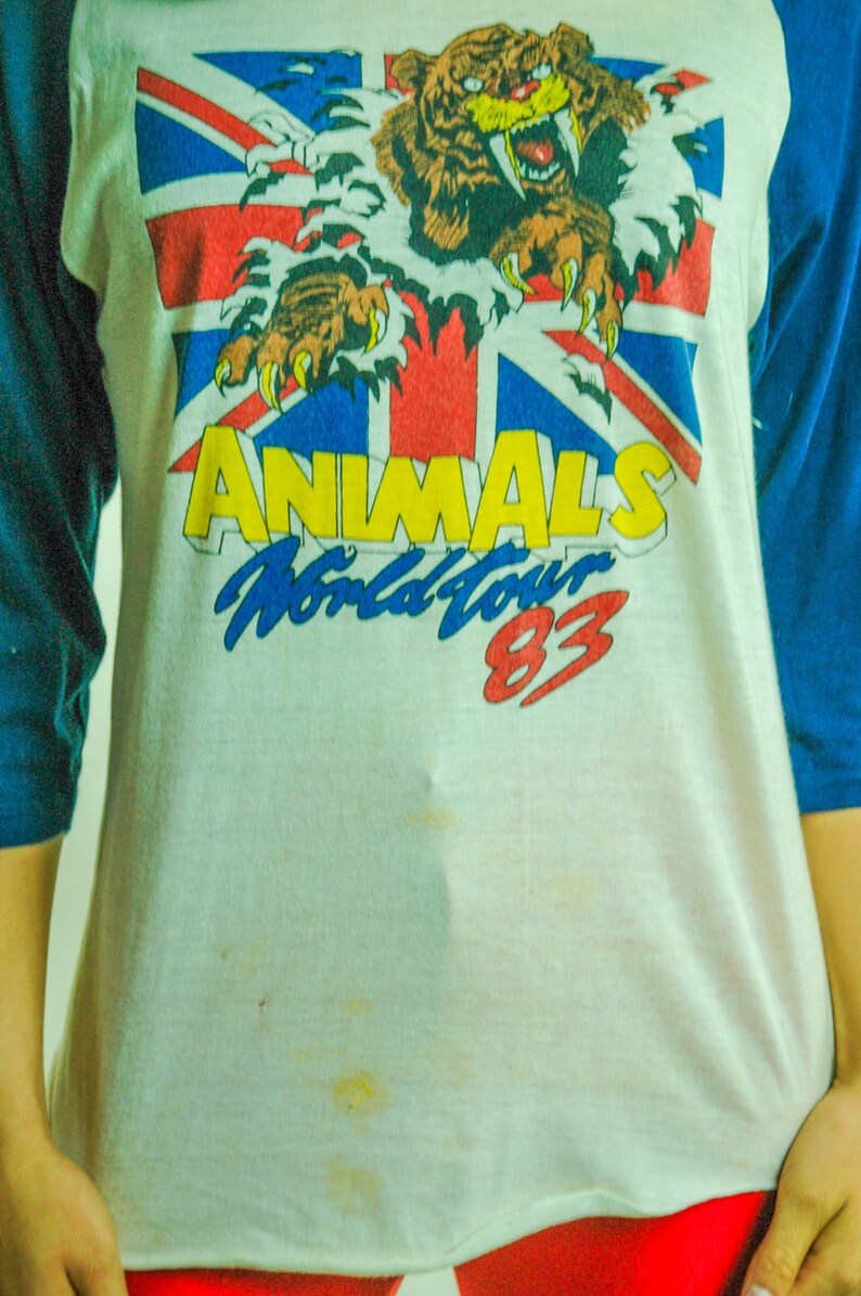 May include: A white t-shirt with a blue and red Union Jack flag design. A cartoon tiger with fangs is breaking through the flag. The text "ANIMALS World Tour 83" is printed below the tiger.