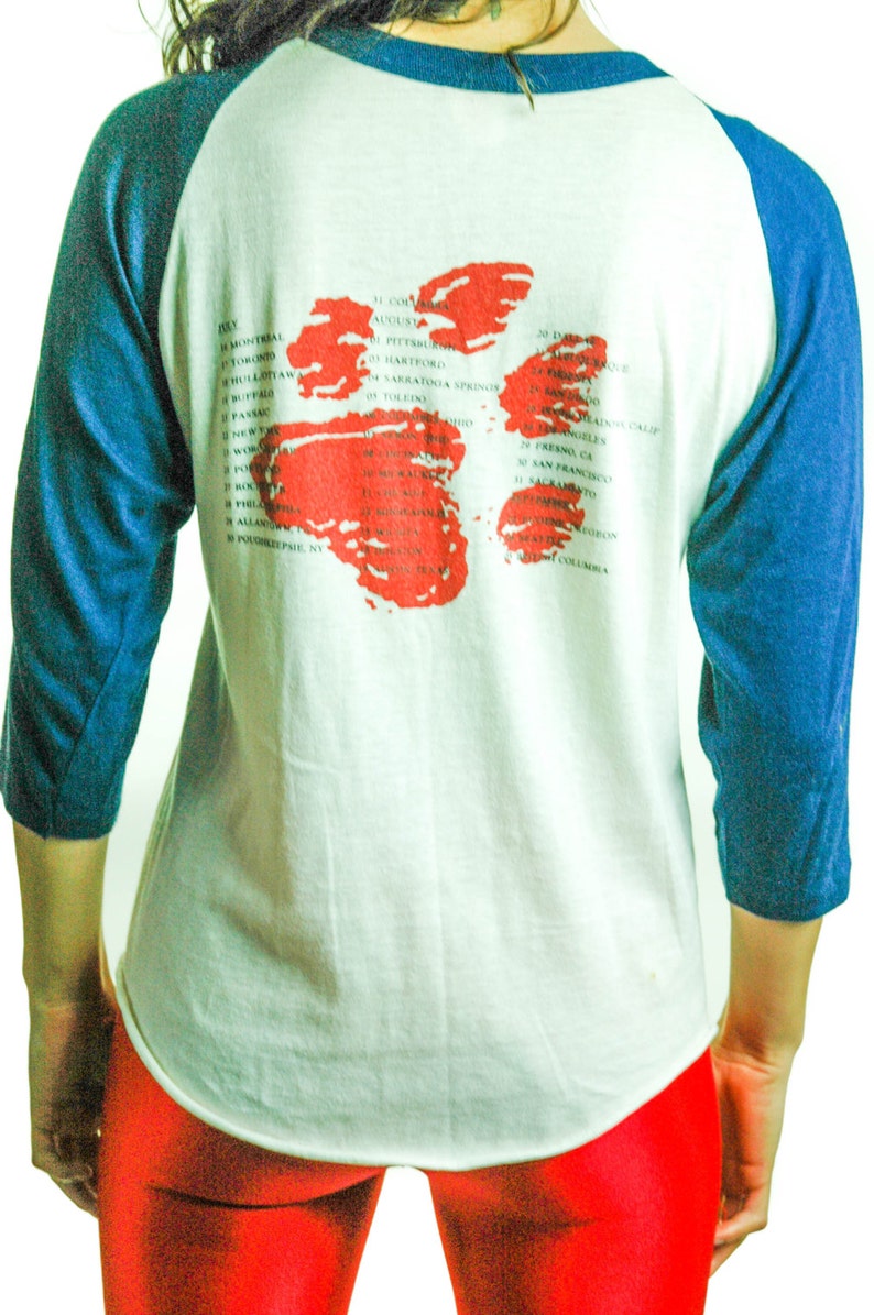 May include: A white and blue baseball style t-shirt with a red splatter design on the back. The shirt has a list of cities and dates printed on the back.
