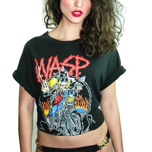 May include: A black t-shirt featuring a graphic of a cartoon character riding a motorcycle with flames. The text "WASP" is printed in red on the shirt. The shirt is cropped and worn with a black bikini bottom with a gold buckle.