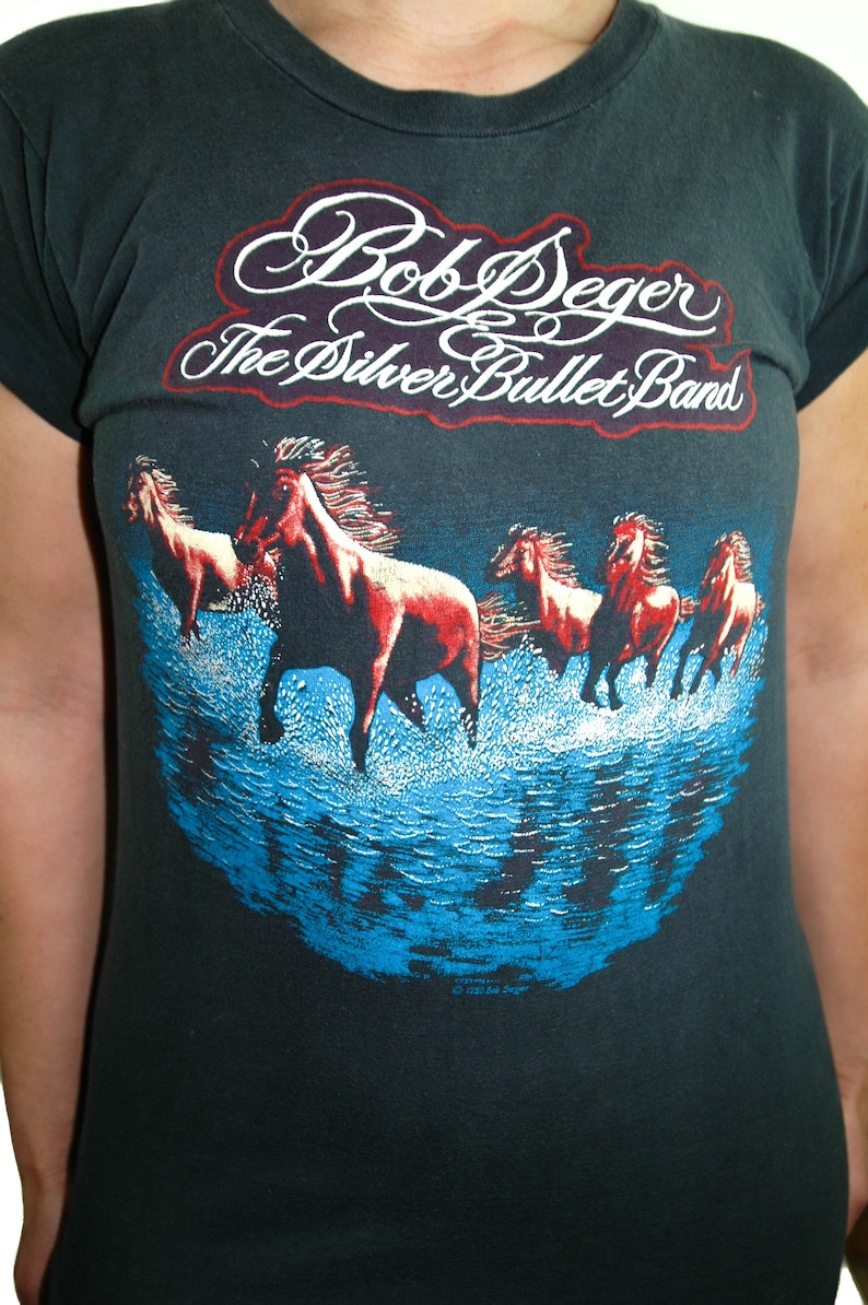May include: A black t-shirt with a white graphic print of Bob Seger and The Silver Bullet Band. The graphic features five horses running through water.