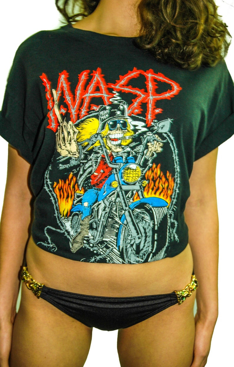 May include: A black t-shirt featuring a graphic of a skeleton riding a motorcycle. The graphic includes the word "WASP" in barbed wire. The t-shirt is being worn by a person with long brown hair.