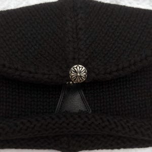May include: A black knit hat with a silver-colored button clasp. The hat features a textured knit pattern and a small, triangular leather detail beneath the button. The button has a decorative floral design.