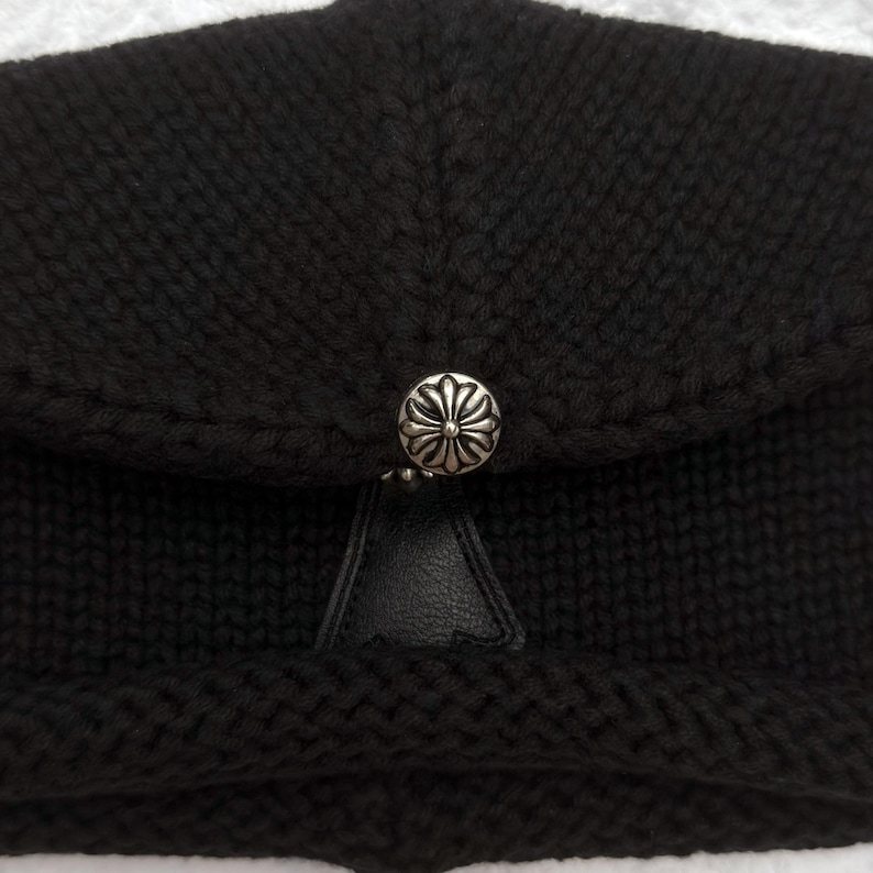 May include: A black knit hat with a silver-colored button clasp. The hat features a textured knit pattern and a small, triangular leather detail beneath the button. The button has a decorative floral design.