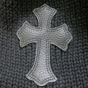 Puede incluir: Aplicaci&oacute;n de cruz de cuero negro con superficie texturizada cosida sobre tejido de punto gris oscuro. La cruz tiene un dise&ntilde;o decorativo y ornamentado con bordes redondeados y est&aacute; centrada.