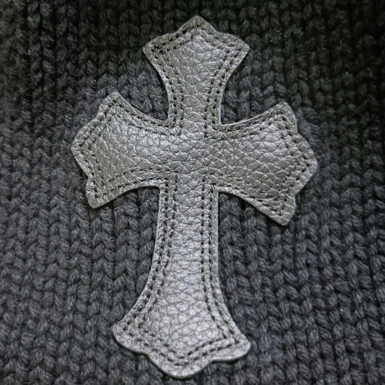 Puede incluir: Aplicaci&oacute;n de cruz de cuero negro con superficie texturizada cosida sobre tejido de punto gris oscuro. La cruz tiene un dise&ntilde;o decorativo y ornamentado con bordes redondeados y est&aacute; centrada.
