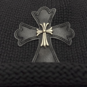 May include: A dark gray knit beanie featuring a black leather cross applique with a silver-colored center. The cross is detailed with decorative stitching. The beanie has a ribbed knit texture.