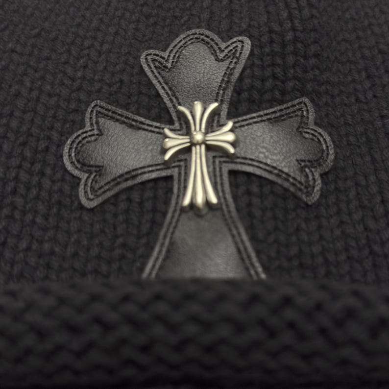 May include: A dark gray knit beanie featuring a black leather cross applique with a silver-colored center. The cross is detailed with decorative stitching. The beanie has a ribbed knit texture.