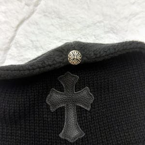 Puede incluir: Gorro de punto negro con una aplicaci&oacute;n de cruz de cuero y un bot&oacute;n decorativo plateado. El gorro tiene un borde doblado y una apariencia texturizada. La cruz es un elemento de dise&ntilde;o prominente.