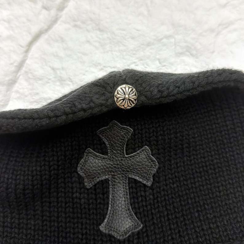 Puede incluir: Gorro de punto negro con una aplicaci&oacute;n de cruz de cuero y un bot&oacute;n decorativo plateado. El gorro tiene un borde doblado y una apariencia texturizada. La cruz es un elemento de dise&ntilde;o prominente.