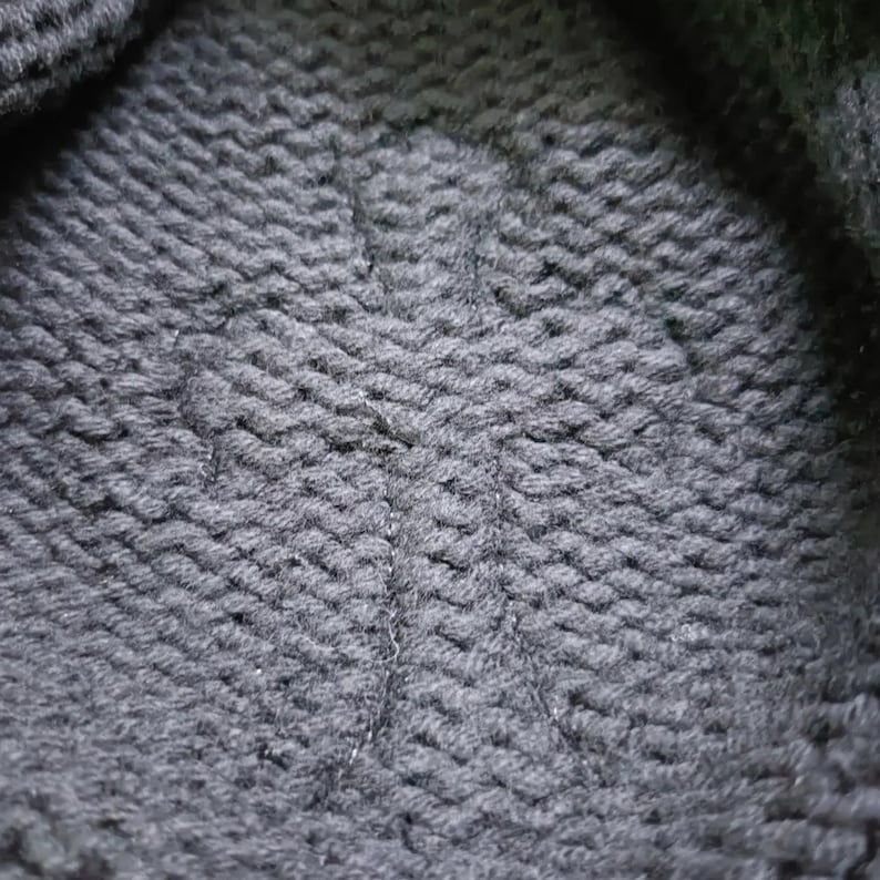 Puede incluir: Primer plano de una tela de punto gris oscuro con un patr&oacute;n texturizado y tejido. La tela parece gruesa y c&aacute;lida, con puntadas visibles que crean un efecto de nervaduras diagonales. El material es probablemente lana o una fibra similar.