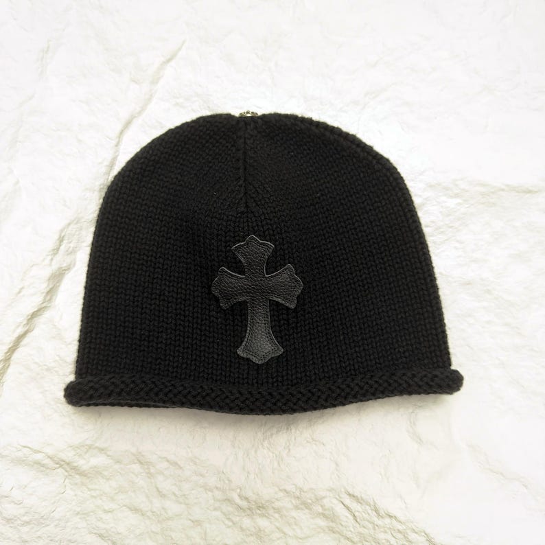 Puede incluir: Un gorro negro de punto con un aplique de cruz de cuero. El gorro tiene un borde doblado y un aspecto texturizado. La cruz est&aacute; centrada en la parte delantera del gorro.