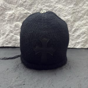 Könnte beinhalten: Schwarze Beanie-Mütze mit einem Kreuzemblem auf der Vorderseite und einer kleinen silberfarbenen Verzierung oben. Die Mütze ist aus einem strukturierten Stoff gefertigt.