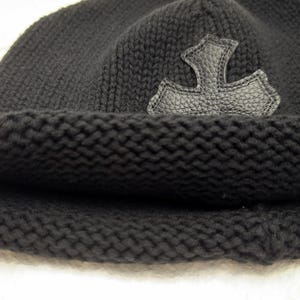 Puede incluir: Gorro de punto negro con borde doblado. Una aplicaci&oacute;n de cuero en forma de cruz estilizada est&aacute; cosida en el lateral. El gorro est&aacute; hecho de un material de punto grueso y texturizado.