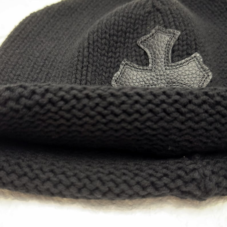 Puede incluir: Gorro de punto negro con borde doblado. Una aplicaci&oacute;n de cuero en forma de cruz estilizada est&aacute; cosida en el lateral. El gorro est&aacute; hecho de un material de punto grueso y texturizado.