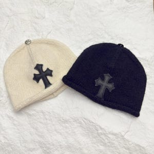 May include: Two knitted beanies, one cream-colored and one black, each featuring a black cross applique. The cream beanie has a silver embellishment on top. The beanies are displayed on a textured, off-white surface.