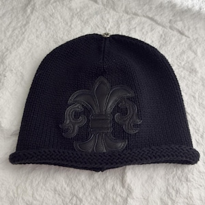 Op de afbeelding: Een marineblauwe gebreide beanie met een zwart leren fleur-de-lis applicatie op de voorkant. De beanie heeft een omgeslagen rand en een kleine zilverkleurige versiering aan de bovenkant. De hoed is gemaakt van een dik, getextureerd breiwerk.