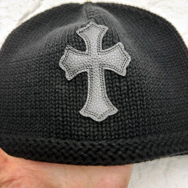 Puede incluir: Gorro de punto negro con una cruz de cuero gris. El gorro tiene un borde acanalado y un patr&oacute;n de punto texturizado. La cruz est&aacute; cosida en el sombrero, agregando un elemento decorativo al tocado.