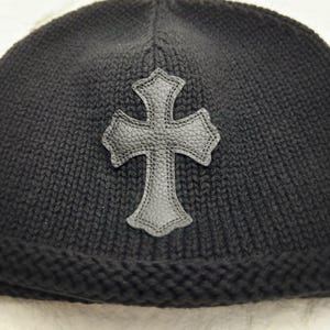 Puede incluir: Gorro de punto negro con una cruz de cuero. El gorro est&aacute; hecho de un material de punto grueso y texturizado. La cruz es de color gris oscuro y tiene una superficie texturizada.